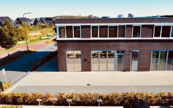 Exterior view of the office building at Deltazijde 20b, Blaricum, featuring a modern brick facade with large windows and a paved entrance. Ideal office space rental option with ample natural light.