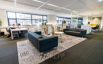 Spacious and modern office space in Breda Centrum-Station, with workstations, a seating area, and large windows. Ideal for those looking to rent office space. A person is working at a desk.