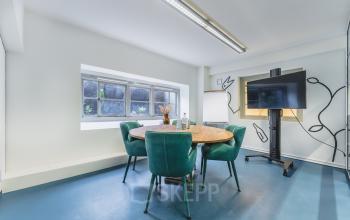 Bright office space in Breda Centrum-Station, featuring a round wooden table with green chairs, a flat-screen TV, and a whiteboard, ideal for office space rental.