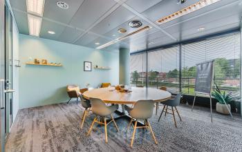 Modern conference room with round table, six chairs, and large windows offering natural light, located at Heerbaan 14, Breda Oost-Brabantpark. Ideal office to rent for collaborative meetings.