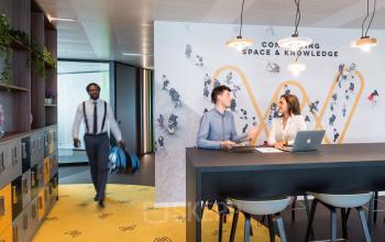 Modern office workspace at Leonardo Da Vincilaan 1, Brussels Airport, with people collaborating at a high table and lockers along the wall.