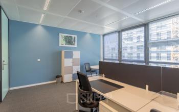 Modern office space with large windows, blue walls, desks, chairs, and storage cabinets at Meeûssquare 37, Brussels European District, Brussels. Ideal for office space rental.