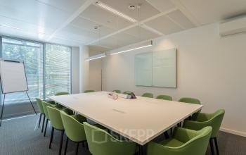 Modern conference room in Brussels European District, located at Meeûssquare / Square de Meeûs 37, with a large table, green chairs, a whiteboard, and natural light through large windows, ideal for office space rental.