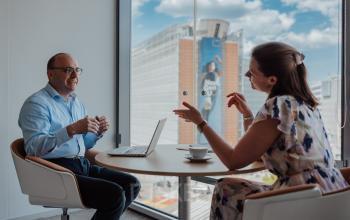 Two business professionals engage in a discussion at a round table in a modern office with a view of Brussels European District, ideal for office space rental.