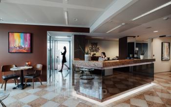 Modern office with a reception desk and a small meeting area. A person works at the desk while another walks by, set in Brussels European District.
