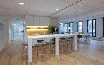 Modern office pantry at Willemsplein 2, Den Bosch, featuring a kitchen area with bar stools and tables, available for office space rental.