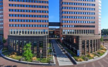 Exterior view of the office building at Utopialaan 22, Den Bosch, showcasing a modern architectural design with large windows and a spacious pathway entrance. Ideal for office space rental opportunities.