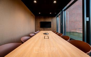 Modern conference room at Hambakenwetering 5 with a long wooden table, surrounded by chairs and a wall-mounted screen, ideal for office space rental in Den Bosch.