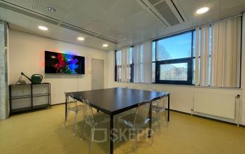 Bright and modern conference room at Bruistensingel 500, Den Bosch, featuring a large table with transparent chairs, ample natural light from windows, and vibrant wall art, ideal for office space rental.