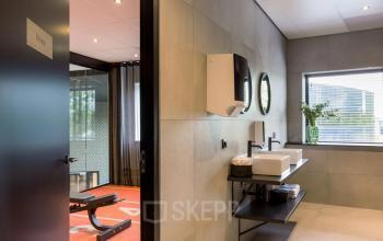 Modern office space interior at Reitschweg 1, Den Bosch, featuring a sleek restroom with a dual sink and a glimpse of a workout area. Ideal for office space rental.