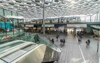 Spacious interior of Koningin Julianaplein 10 in The Hague Center featuring people engaging in various activities. A train passes through the office space rental area.