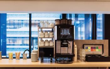 Office pantry area with a modern coffee machine, cups, and accessories set against a backdrop of large windows in The Hague Center. Ideal for office space rental.