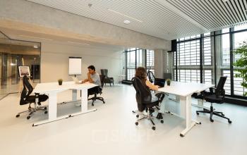 Bright office space with large windows, featuring desks and chairs, located at Waldopstraat 5. Two individuals are engaged in work activities, ideal for an office space rental in The Hague Center.