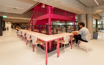 Modern office space rental at Waldopstraat 5, The Hague Center, with people working at communal desks near a vibrant staircase.