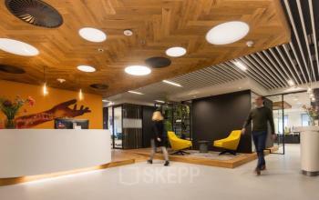 Reception area at Prinses Margrietplantsoen 25, The Hague Haagse Hout, with modern lighting, wooden ceiling, and yellow chairs, ideal for office space rental. People are engaged in movement.