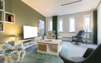Interior of an office at Keulenstraat 12 in Deventer with comfortable seating, a TV, bookshelves, and ample light from windows.