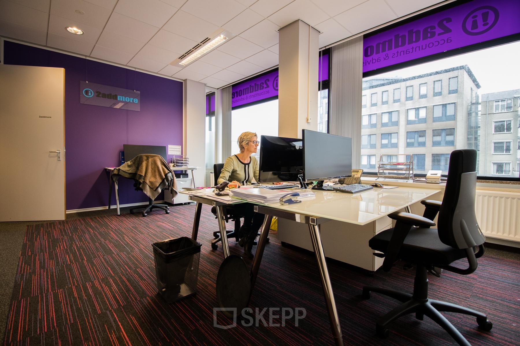 Modern office space with large windows at Berglaan 4-8, Drachten, featuring workstations, chairs, and a business professional working at a desk.
