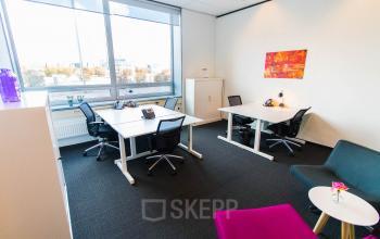 A bright office space for rent at Eindhoven Airport with four desks, ergonomic chairs, and a colorful wall art, offering a functional workspace for productivity.