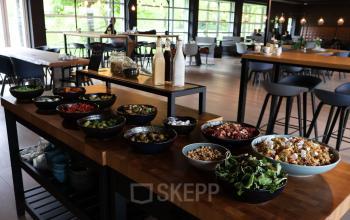 A spacious, well-lit lounge area at Flight Forum 760-780, Eindhoven, with a stylish buffet setup featuring a variety of fresh salads and condiments. Ideal for networking in a casual office space rental.