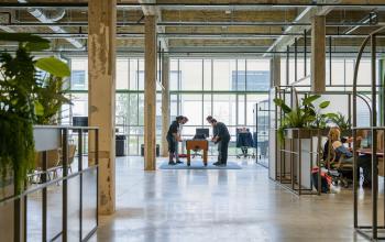 Spacious office to rent at Frederiklaan 10A, Eindhoven Strijp-S with industrial design, featuring open workspaces, natural light, and people collaborating at a meeting table amidst modern furnishings.