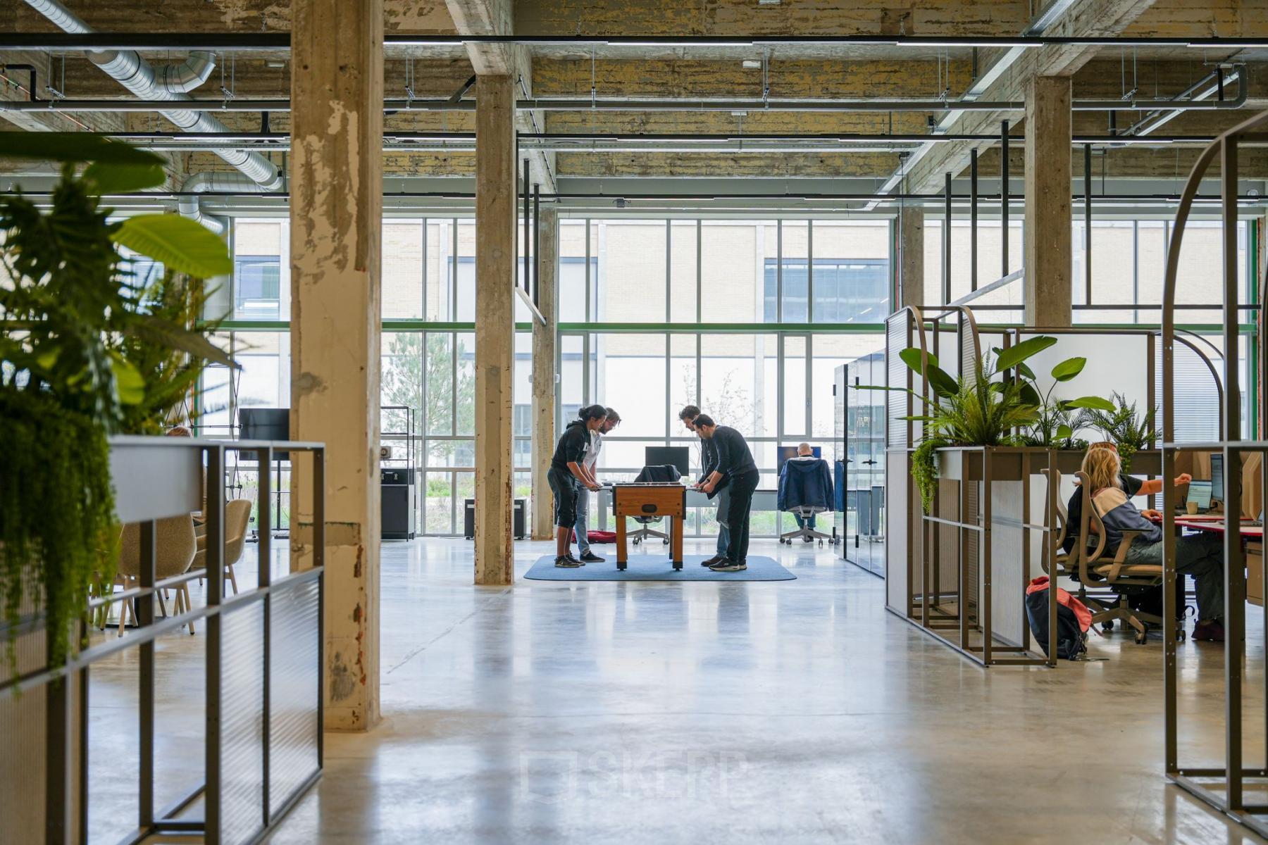 Spacious office to rent at Frederiklaan 10A, Eindhoven Strijp-S with industrial design, featuring open workspaces, natural light, and people collaborating at a meeting table amidst modern furnishings.