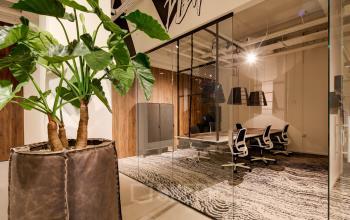Modern office space available for rent at Frederiklaan 10E, Eindhoven Strijp-S, featuring a stylish meeting area with glass walls, wooden accents, and a large plant for ambience.