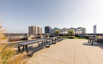 Spacious rooftop terrace at Brouwerijstraat 1, Enschede, with picnic tables, planters, and a view of the cityscape.