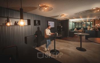 Modern office lounge at Brouwerijstraat 1, Enschede, featuring stylish lighting, high tables, and professionals engaged in business activities. Ideal office space rental for collaborative environments and networking.