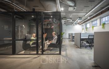 Modern office space rental at Brouwerijstraat 1, Enschede featuring an open floor plan with a stylish glass meeting booth, industrial decor, and contemporary furniture ideal for productivity.