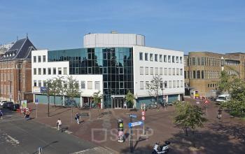 Street view of the office building at Hoedemakerplein 2, Enschede, showcasing a modern exterior with large windows. Ideal for office space rental, the building stands prominently in a bustling urban area.