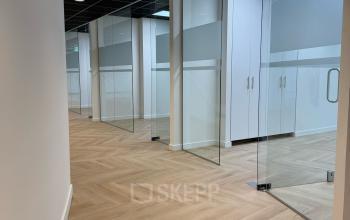 Modern office hallway at Hoedemakerplein 2 in Enschede, featuring light wood flooring and glass doors leading to private office spaces available for rent.