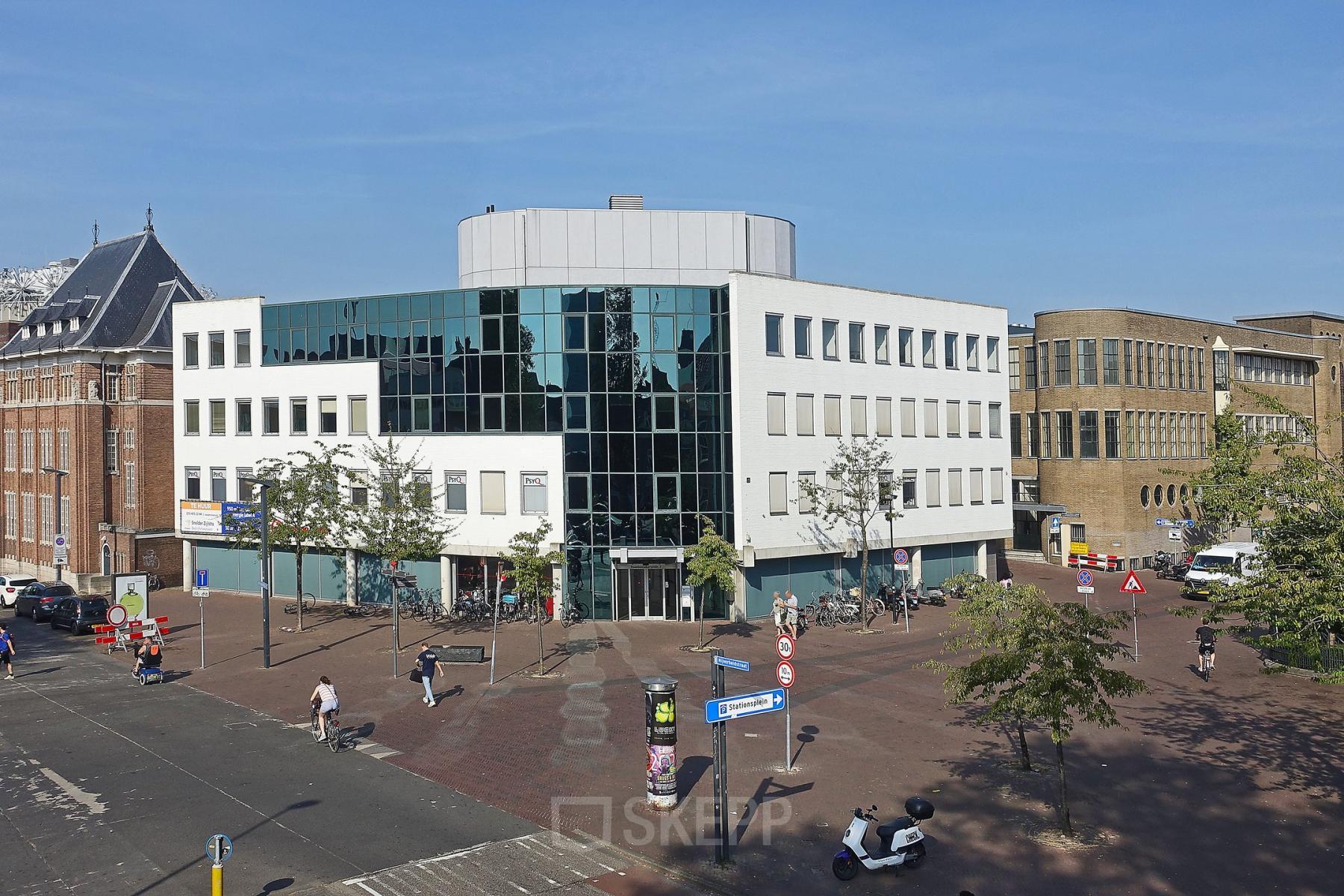 Street view of the office building at Hoedemakerplein 2, Enschede, showcasing a modern exterior with large windows. Ideal for office space rental, the building stands prominently in a bustling urban area.