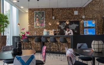 Moderner, möblierter Büroraum in der Baseler Straße 10, Frankfurt Hauptbahnhof. Barbereich mit hängenden Lampen, dekorativen Stühlen und einer Person bei der Arbeit am Tresen.