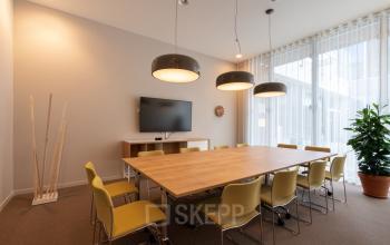 Vergaderruimte met grote houten tafel, gele stoelen, drie hanglampen, tv-scherm en witte gordijnen in het kantoor te huur aan Gaston Crommenlaan 8, Gent Ledeberg, Gent.