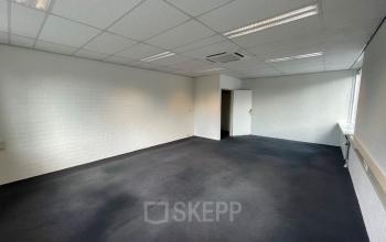 Interior of an office space for rent at Techniekweg 15, Gorinchem, with black carpet flooring, white walls, overhead lighting, and a door leading to another room.