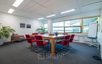 Bright office space rental in Gouda with a spacious conference table and red chairs, complemented by large windows and green plants.