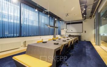 Modern conference room at Kennemerplein 6-14 in Haarlem Centraal Station, Haarlem, featuring a large table, sleek chairs, blue carpet, ceiling lights, and a presentation screen, available for office space rental.