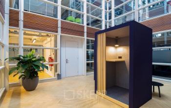 Modern work booth with wooden floors at Kennemerplein 6-14 office space rental in Haarlem Centraal Station, surrounded by potted plants and windowed walls.
