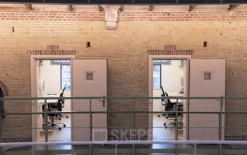 Interior view of office space rental at Koepelplein 1E in Haarlem Centrum, featuring two separate workspaces with desks, chairs, and visible computers, against a backdrop of exposed brick walls.