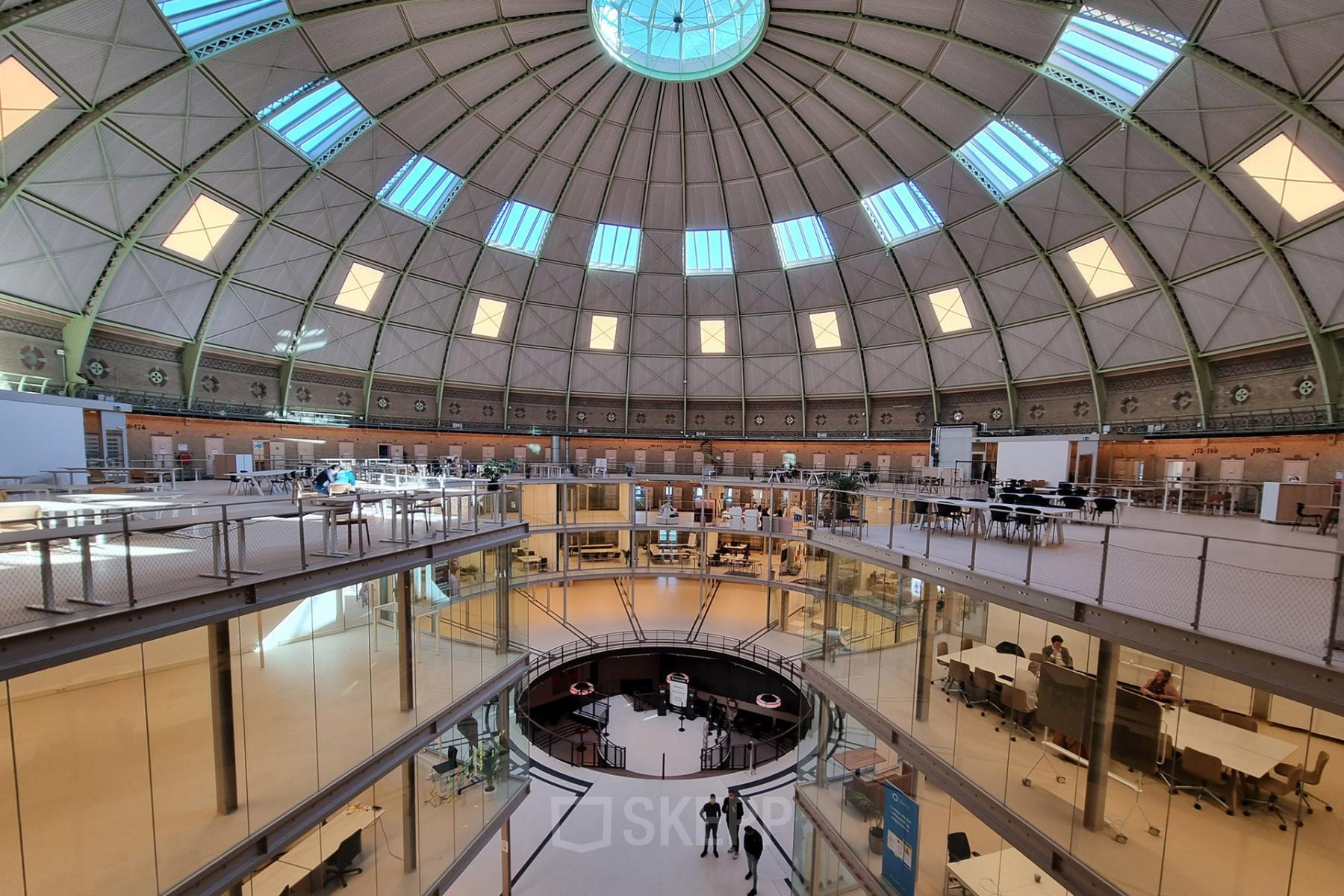Spacious office to rent at Koepelplein 1E, Haarlem Centrum. Featuring a dome ceiling with abundant natural light, furnished workspaces and people engaged in discussions, ideal for office space rental.