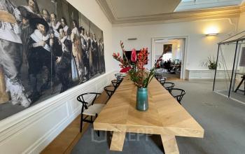 Modern conference room with a long wooden table, surrounded by black chairs, large painting on the wall, and a vase with flowers at Nieuwe Gracht 74-76, Haarlem Centrum, available for office space rental.