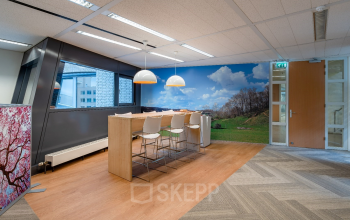 Modern office space in Heerlen at Geerstraat 115 with a high table and bar stools, hardwood floors, and a vibrant wall mural. Perfect for office space rental with natural light streaming through large windows.