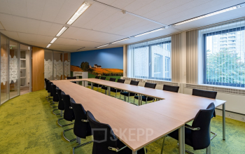 Spacious conference room with a large U-shaped table and modern chairs, ideal for office space rental at Geerstraat 115, Heerlen. The room features natural lighting and a scenic mural.