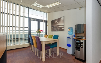 Office space rental at Milaanstraat 70-110, Heerlen, featuring a compact meeting area with colorful chairs, a wooden table, modern water dispenser, and a coffee machine, creating a welcoming work environment.