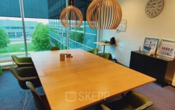 Bright office space rental in Snellius 1, Heerlen with a large wooden table, green chairs, pendant lights, and a view through floor-to-ceiling windows.