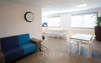Bright office space at Steenovenweg 5, Helmond, featuring a comfortable seating area with a clock on the wall, and a larger table with chairs next to a window.