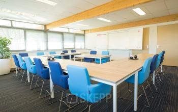 Modern conference room at Steenovenweg 5, Helmond, featuring a large table surrounded by blue chairs, whiteboard, and ample lighting from large windows.
