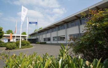 Exterior view of the office building located at Steenovenweg 5, Helmond, with multiple company flags in front and greenery surrounding the entrance area.
