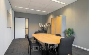 A modest conference room with a wooden table and black chairs, featuring a cabinet and decorative plants, ideal for office space rental.