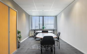 Bright office space for rent in Hengelo, featuring a meeting table with chairs, large window, potted plants, and built-in storage, creating a functional workspace atmosphere.
